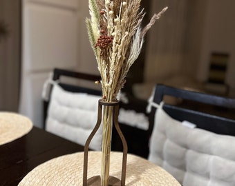 May include: A wooden vase with a clear glass tube insert, holding a bouquet of dried pampas grass, white and brown dried flowers, and small brown berries. The vase is sitting on a round, woven placemat.