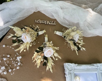 May include: Three small bouquets of dried flowers with white roses and pampas grass. The bouquets are tied with white ribbon and are arranged on a brown surface.