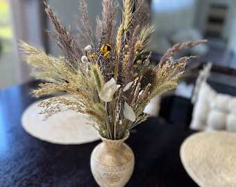 Può includere: Un arrangiamento di fiori secchi in un vaso di ceramica beige con un motivo a chiave greca. Il vaso è appoggiato su un tavolo di legno con un sottopentola intrecciato.