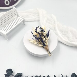 Op de afbeelding: Een klein boeket gedroogde bloemen in tinten wit en paars, gebonden met touw, op een witte ronde basis. Het boeket bestaat uit gedroogde grassen, lavendel en kleine witte bloemen.