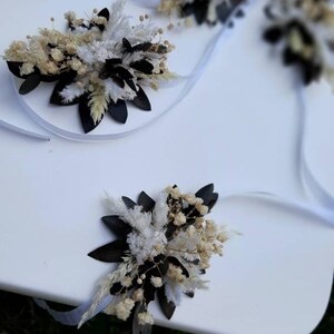 May include: Three white satin ribbons with dried flower arrangements attached. The arrangements feature white and cream dried flowers, white fluffy dried grasses, and black dried leaves.