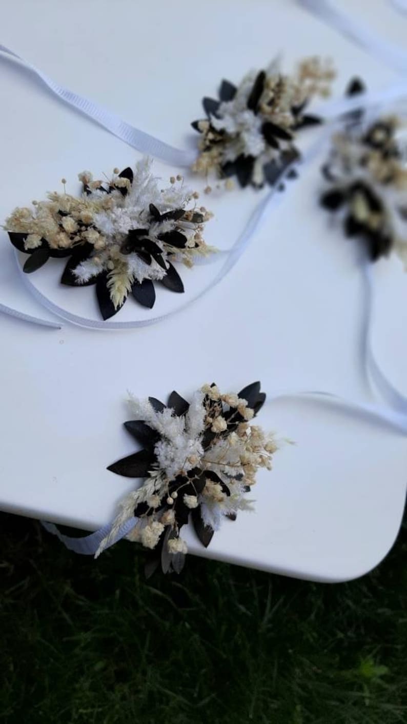 May include: Three white satin ribbons with dried flower arrangements attached. The arrangements feature white and cream dried flowers, white fluffy dried grasses, and black dried leaves.