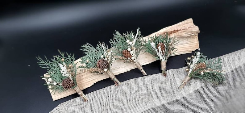 May include: Five small bouquets of greenery, pine cones, and white berries arranged on a piece of wood. The bouquets are tied with twine and are ready to be used as boutonnieres.