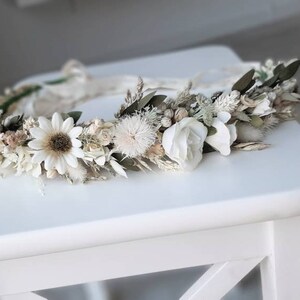 May include: A white floral crown with dried flowers and greenery. The crown is made with white daisies, white roses, and other white flowers. The crown is on a white table.