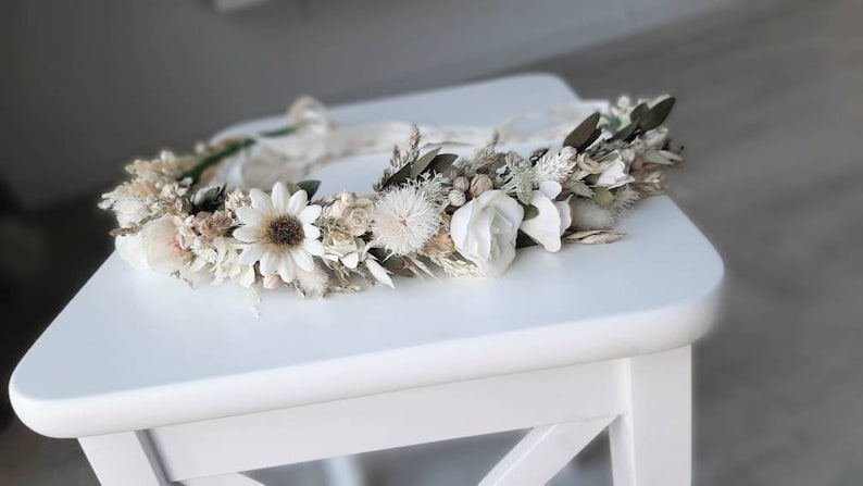 May include: A white floral crown with dried flowers and greenery. The crown is made with white daisies, white roses, and other white flowers. The crown is on a white table.
