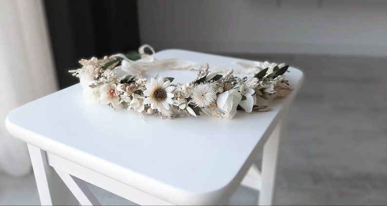 May include: A white and cream floral crown with dried flowers and greenery. The crown is made with a variety of flowers, including daisies, baby's breath, and eucalyptus.