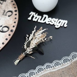 May include: A small bouquet of dried flowers with white and beige blooms and green leaves, tied with twine. The bouquet is on a black surface with a white lace trim and a wooden sign that says "TnDesign".