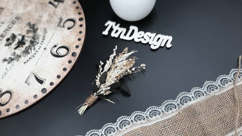 May include: A small bouquet of dried flowers with white and beige blooms and green leaves, tied with twine. The bouquet is on a black surface with a white lace trim and a wooden sign that says "TnDesign".