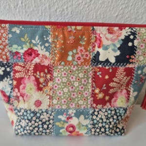 Könnte beinhalten: Eine Patchwork-Kosmetiktasche mit rotem Reißverschluss und kleinem Quastenanhänger. Die Tasche besteht aus Stoffquadraten in verschiedenen Blumenmustern, darunter Rot, Blau, Orange und Grün. Die Tasche hat eine Trapezform.
