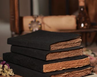May include: A stack of four rectangular journals with matte black covers and thick, textured, light brown deckled-edge paper. The books are arranged in a vertical pile, showcasing the rough, uneven edges of the pages against the dark covers.