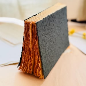 May include: A small book with a textured black cover and thick, uneven, light brown paper pages. The book is standing upright on its edge, showcasing the rough, deckled edges of the paper and the dark, grainy surface of the cover.
