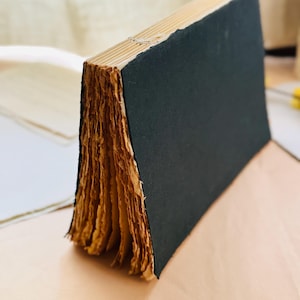May include: A handmade book with a solid black cover and exposed stitching along the spine. The pages are made of thick, textured, light brown paper with rough, deckled edges that are visible from the side of the book.