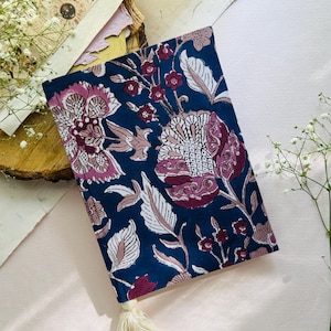 May include: A navy blue fabric journal cover with a floral pattern in shades of pink, white, and burgundy. The design features large stylized flowers and leaves. A cream-colored tassel hangs from the bottom edge.