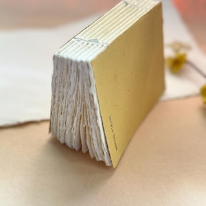 Peut inclure: Un petit carnet avec une couverture jaune moutarde et des pages en papier blanc aux bords irréguliers. La reliure présente des coutures en fil blanc visibles. Le texte "Deckle Dreams" est imprimé en noir sur la couverture.