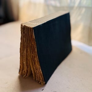 Könnte beinhalten: Ein Buch mit einem einfarbigen schwarzen Einband und sichtbarer Fadenheftung am Buchrücken. Die Seiten sind dick, strukturiert und haben ein hellbraunes, gealtertes Aussehen mit rauen, unebenen Kanten an der Seite.