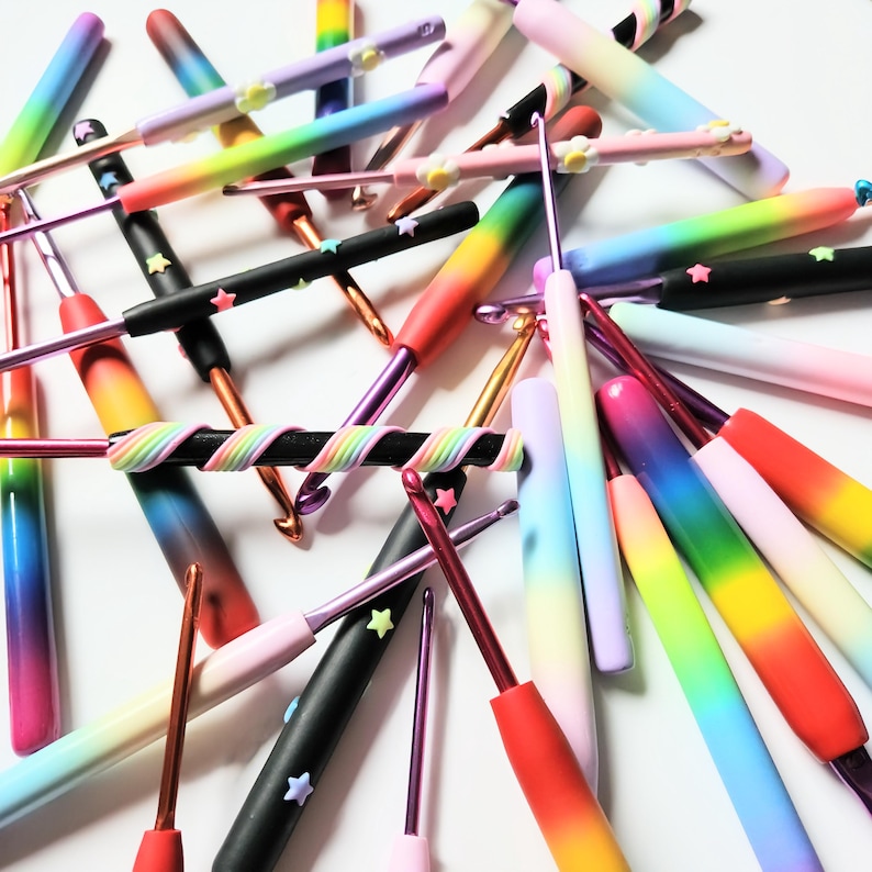 May include: A collection of colorful crochet hooks with rainbow ombre and star accents. The hooks are made of plastic and are arranged in a random pattern on a white surface.