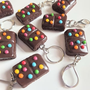 May include: A collection of rectangular chocolate bar keychains featuring a smooth dark brown finish topped with small, colorful candy-like dots. Each piece is attached to a silver-toned metal split ring or a lobster clasp for secure attachment.