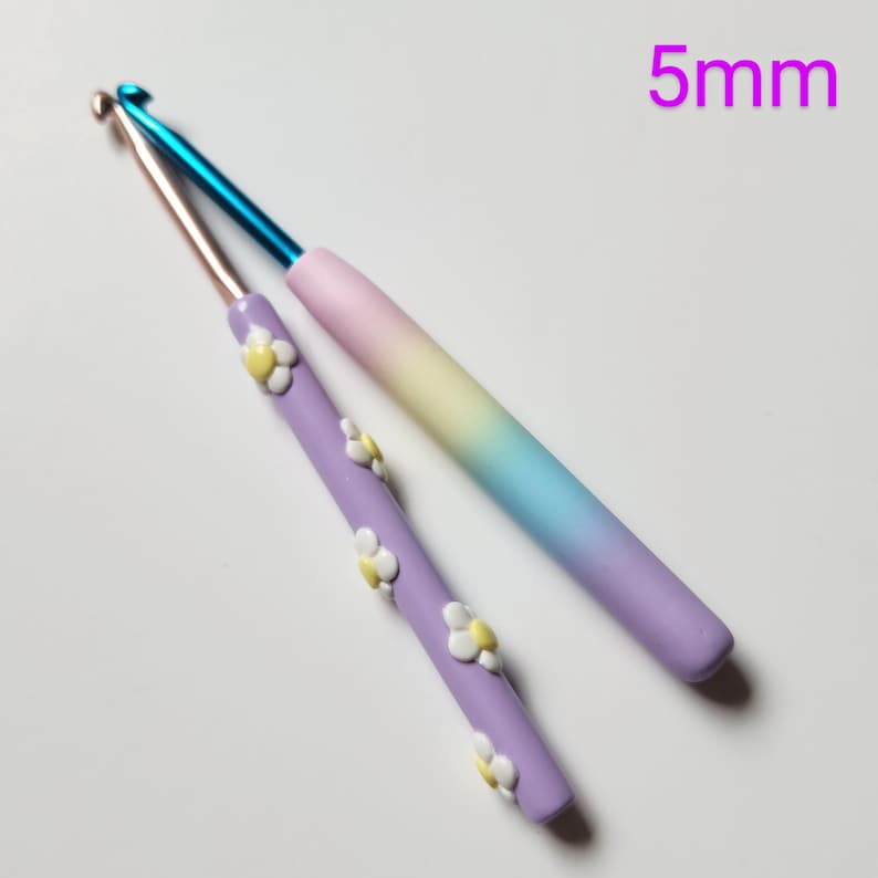 May include: Two crochet hooks, one with a rainbow gradient and the other with a purple base and white daisy accents. Both hooks are 5mm.