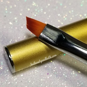 May include: A gold-colored nail brush with a slanted, orange-colored bristle brush head. The brush is labeled "knibeauityandbath.com".