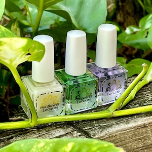 May include: Three bottles of Sunflower Lacquer nail polish in pastel colors: cream, green, and lavender. Each bottle has a white cap and a label with a sunflower design. The bottles are arranged on a wooden surface, surrounded by green leaves.