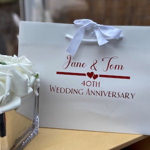 May include: White gift bag with a red ribbon and the text "Jane & Tom 40th Wedding Anniversary" printed on the front.