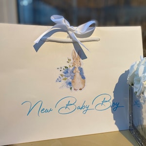 May include: A white gift bag with a white satin ribbon and a blue and white Peter Rabbit sticker. The bag has the text "New Baby Boy" written in blue cursive.