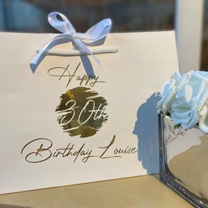 May include: White gift bag with a gold foil design that says "Happy 30th Birthday Louise". The bag is tied with a white ribbon and is next to a clear glass vase filled with white roses.
