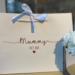 May include: Cream-colored gift bag with the words "Mummy TO BE" in rose gold script and a small heart. The bag has white rope handles and a white satin ribbon tied in a bow at the top. A vase of white roses is in the background.