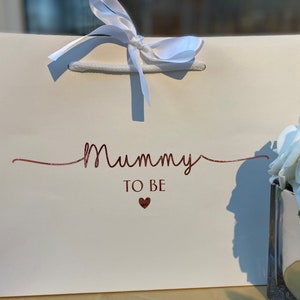 May include: A white gift bag with the words "Mummy TO BE" in a glittery rose gold script. A small heart is below the text. The bag has white rope handles and a white ribbon tied in a bow at the top.