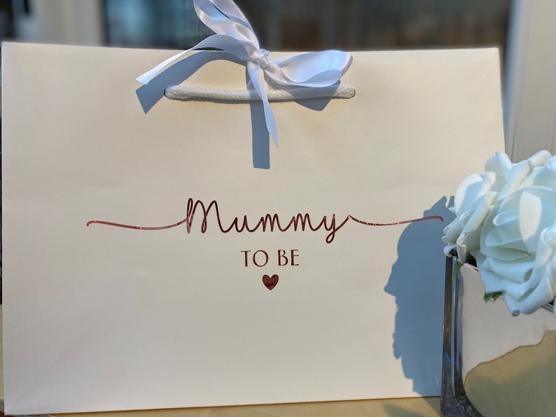 May include: A white gift bag with the words "Mummy TO BE" in a glittery rose gold script. A small heart is below the text. The bag has white rope handles and a white ribbon tied in a bow at the top.