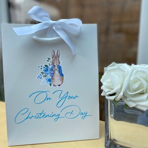 May include: White gift bag featuring a blue and white Peter Rabbit illustration and the text "On Your Christening Day". The bag is tied with a white ribbon.