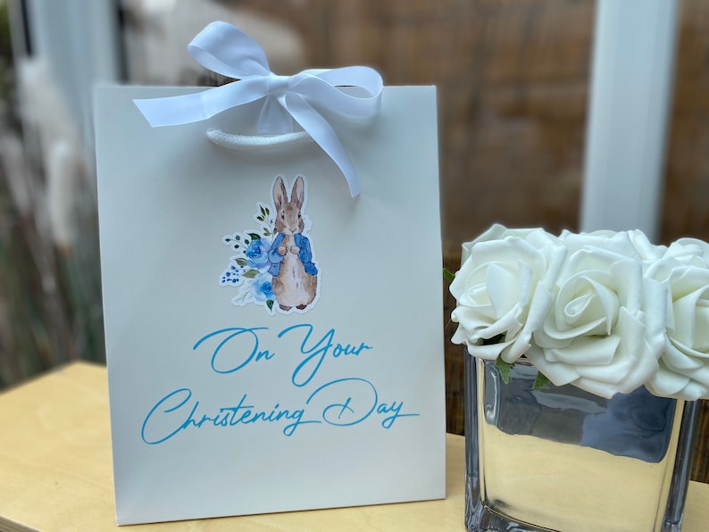 May include: White gift bag featuring a blue and white Peter Rabbit illustration and the text "On Your Christening Day". The bag is tied with a white ribbon.