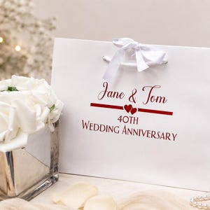 May include: A white gift bag with a white ribbon bow and handles. The bag features the names "Jane & Tom" in red script, with a red line and two hearts, and the text "40th WEDDING ANNIVERSARY" in red.
