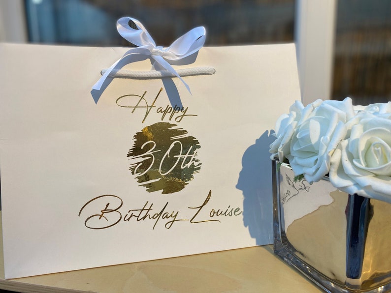 May include: White gift bag with a gold foil design that says "Happy 30th Birthday Louise". The bag is tied with a white ribbon and is next to a clear glass vase filled with white roses.