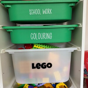 May include: A white shelf unit with four green and white plastic storage bins. The bins are labelled "School Work", "Colouring", and "Lego".