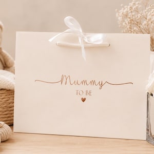 May include: A cream-colored gift bag with a white ribbon and the words "Mummy to be" in a copper-colored script. A small copper heart is below the text. A teddy bear, baby shoes, and a blanket are in the background.