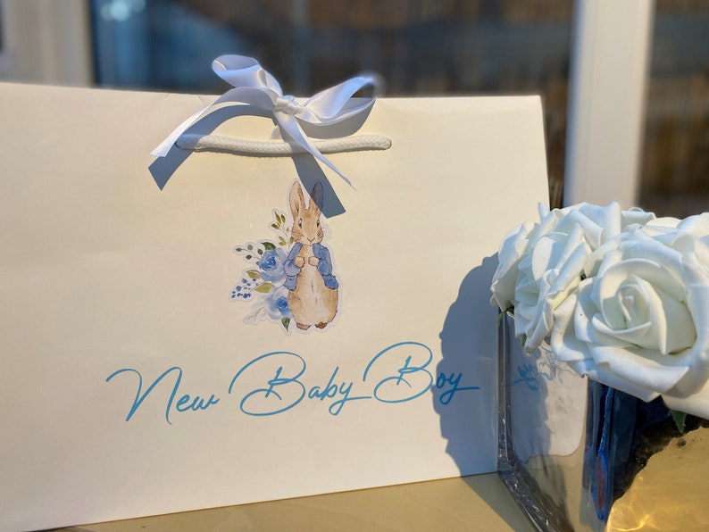May include: A white gift bag with a white satin ribbon and a blue and white Peter Rabbit sticker. The bag has the text "New Baby Boy" written in blue cursive.
