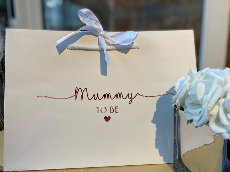 May include: Cream-colored gift bag with the words "Mummy TO BE" in rose gold script and a small heart. The bag has white rope handles and a white satin ribbon tied in a bow at the top. A vase of white roses is in the background.