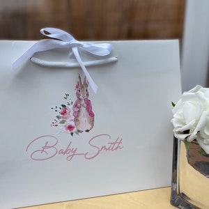 May include: White gift bag with a white ribbon and a pink and brown illustration of a rabbit with flowers. The text "Baby Smith" is written in pink cursive.
