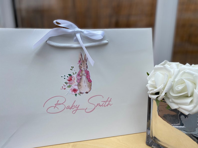 May include: White gift bag with a white ribbon and a pink and brown illustration of a rabbit with flowers. The text "Baby Smith" is written in pink cursive.