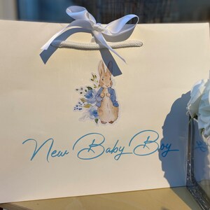 May include: A white gift bag with a white satin ribbon and a blue and white Peter Rabbit sticker. The bag has the text "New Baby Boy" in blue cursive writing.
