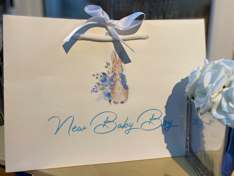 May include: A white gift bag with a white satin ribbon and a blue and white Peter Rabbit sticker. The bag has the text "New Baby Boy" in blue cursive writing.