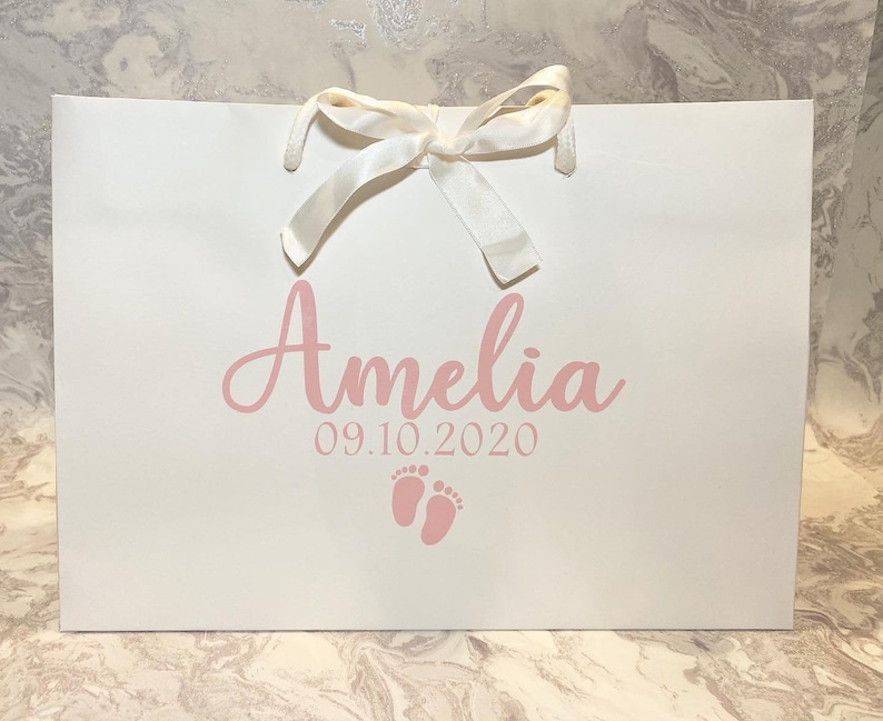 May include: White gift bag with a white satin ribbon and pink lettering. The bag says "Amelia 09.10.2020" and has a pink footprint design.