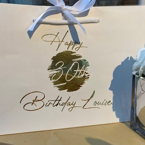 May include: White gift bag with a gold foil design that says "Happy 30th Birthday Louise".