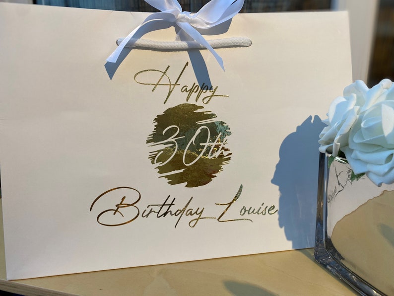 May include: White gift bag with a gold foil design that says "Happy 30th Birthday Louise".