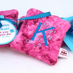 May include: Two pink fabric hand warmers with a blue and white polka dot ribbon and a tag that says "Warm Hands Warm Heart Thank you for all you do! Kate & Kimberly". A third pink hand warmer with a blue embroidered letter K and a blue and white polka dot ribbon. A blue gel ice pack with a white tag.