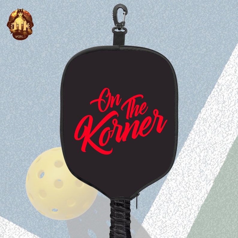 Puede incluir: Raqueta de pickleball negra con un logotipo rojo "On the Korner" y una funda de transporte negra con cremallera. La raqueta tiene un mango de cord&oacute;n negro con un mosquet&oacute;n negro.
