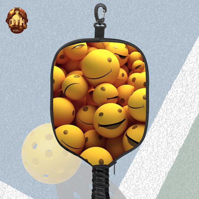 Puede incluir: Una pala de pickleball negra con un dise&ntilde;o de cara sonriente amarilla. La pala tiene un mango negro y un clip negro.