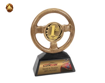 May include: Gold-colored steering wheel trophy with a black base. The base has a gold plaque with the text "LUXCAR Quad A Division Jerry Munson".