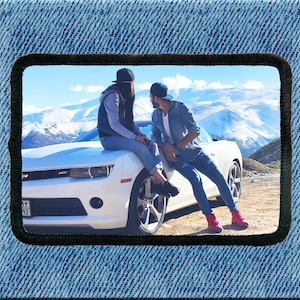 Puede incluir: Un coche deportivo blanco con una pareja sentada en el capó. El coche está aparcado en un camino de tierra con una cordillera nevada al fondo.